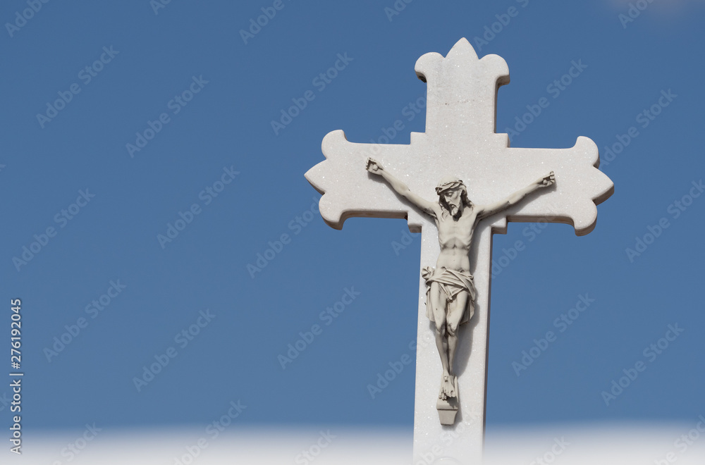 A white cross with a crucified jesus sculpted on it stands against a blue sky background.Thin deliberate blurred white line runs across the bottom of image to add ethereal effect - Image