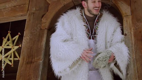 Cheerful young bearded handsome man walking out of the old-fashioned wooden house in national clothes and stylish woolen white coat, acting happily. Good mood, customs and traditions