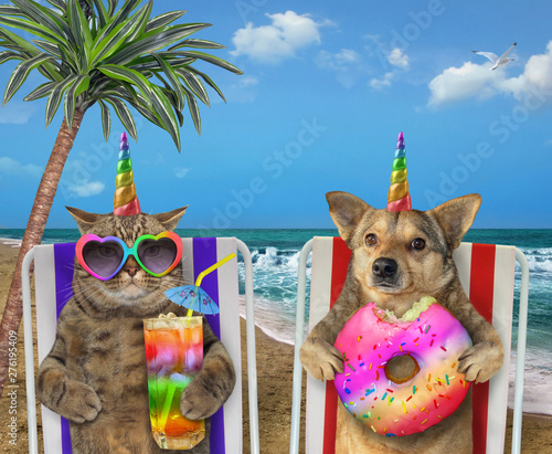 The dog with a donut and the cat with a glass of colored cocktail are sitting under the palm tree on a beach chairs on the beach. © iridi66