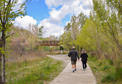 back view of a senior couple walking on a concrete way in the middle of a park. The male hiker wears sorts and a dark blue coat and the female hiker wears black tracksuit and coat. Horizontal picture