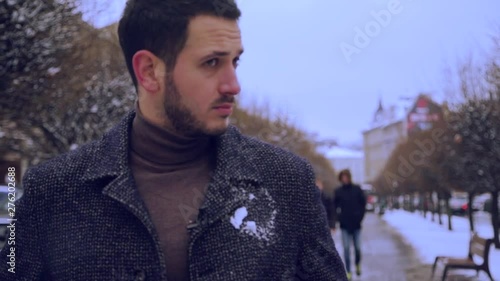 Handsome young man walks down the snowy alley, as he gets a snowball in his body, looks around surprised.Shocking, playful mood, having fun. Romantic atmosphere, winter activities