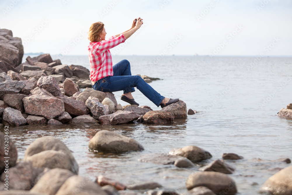 Mature Caucasian woman using smartphone for taking selfie while sitting on rocky seashore, female dressed red and white shirt and blue jeans