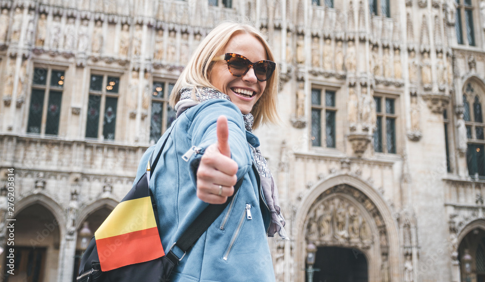 A female traveler with a backpack and a flag of Belgium stands on Grand Place Square in Brussels and shows her thumbs up - Like, Belgium.