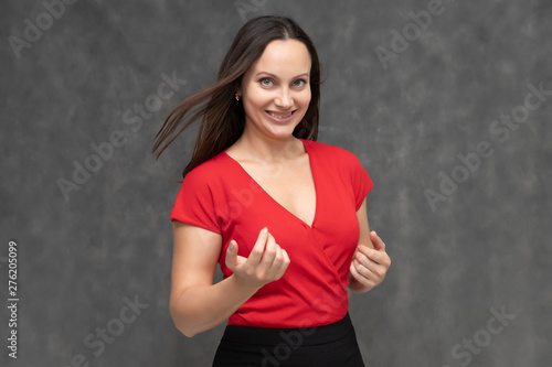 Portrait to the waist of a young pretty brunette woman of 30 years old in a bright red sweater with beautiful dark hair. It is standing on a gray background, talking, showing hands, with emotions