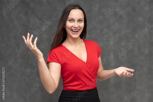 Portrait to the waist of a young pretty brunette woman of 30 years old in a bright red sweater with beautiful dark hair. It is standing on a gray background, talking, showing hands, with emotions