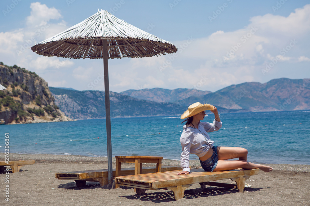young sexy girl in shorts and shirt and hat sitting on a sunbed on the beach by the sea