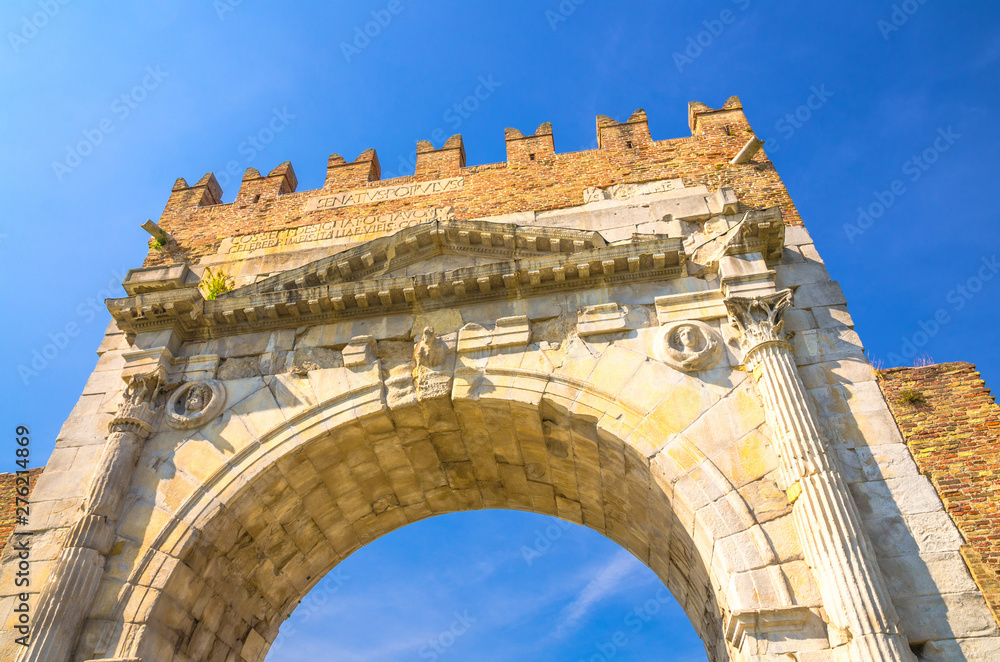 View from below of ancient brick wall and stone gate Arch of Augustus Arco di Augusto ruins in old historical touristic city centre Rimini with blue sky background, Emilia-Romagna, Italy