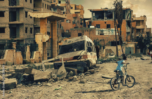 Homeless little boy riding bicycle in destroyed city, military soldiers and helicopters and tanks are still attacking the city photo