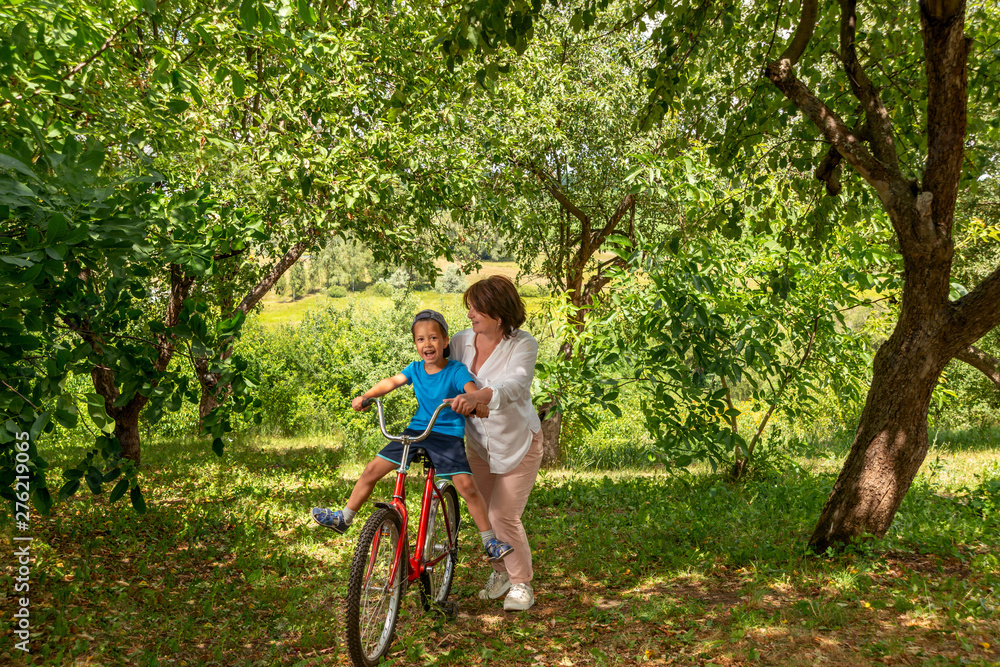 A grandmother  rides on a big red bike of a grandson  in a garden or in a forest on a sunny summer day