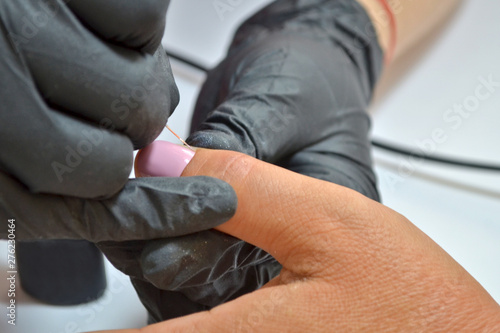 A manicure master in black gloves applies pink nail polish by a thin brush