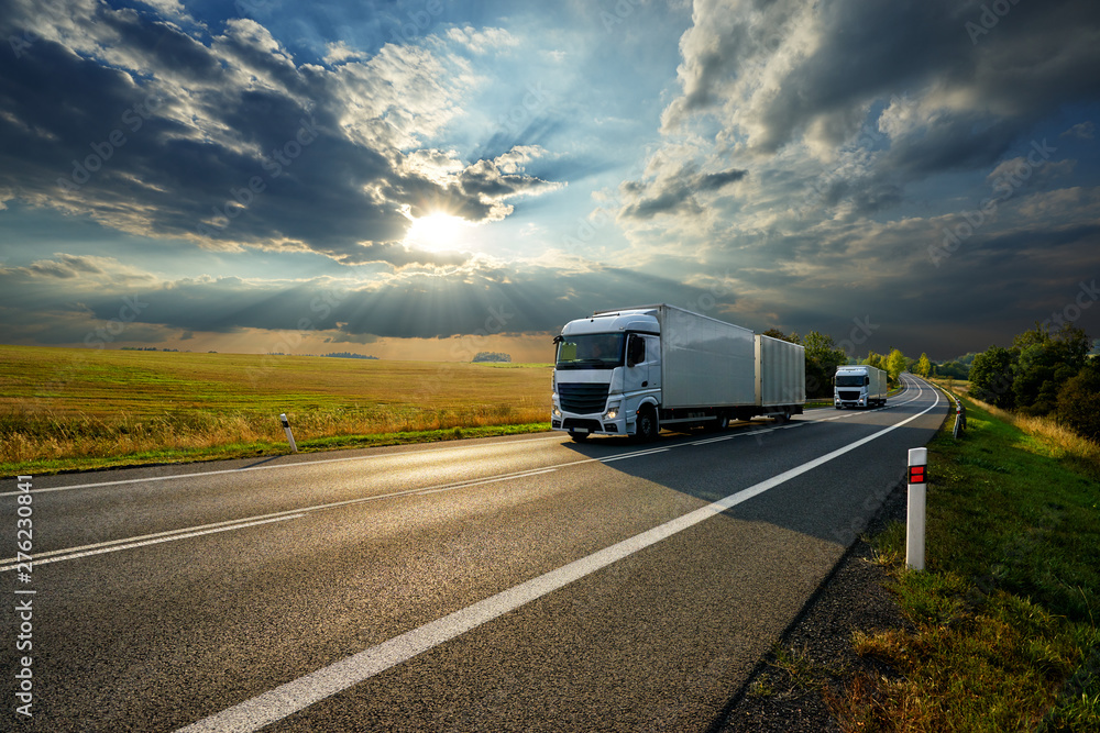 Two white trucks driving on the asphalt road in rural landscape at sunset with dramatic clouds