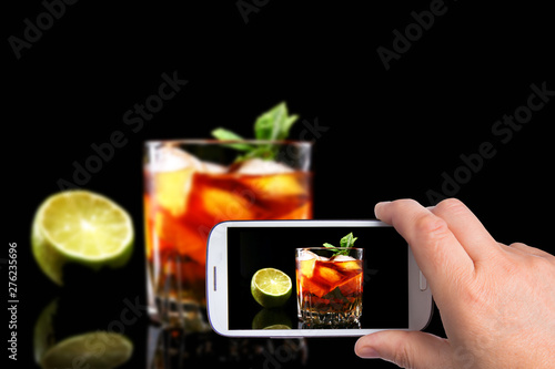hands of man with smartphone taking photo Glass of Dark Rum Cocktail with lime, orange, ice cubes and mint leaves on black mirror background.
