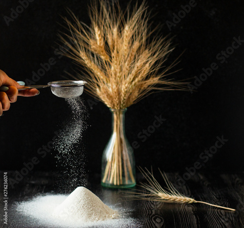 Female hand holds sieve and dissipates perfect flour. Bouquet of the wheat in the vase and handful of the flour and on the dark wooden background