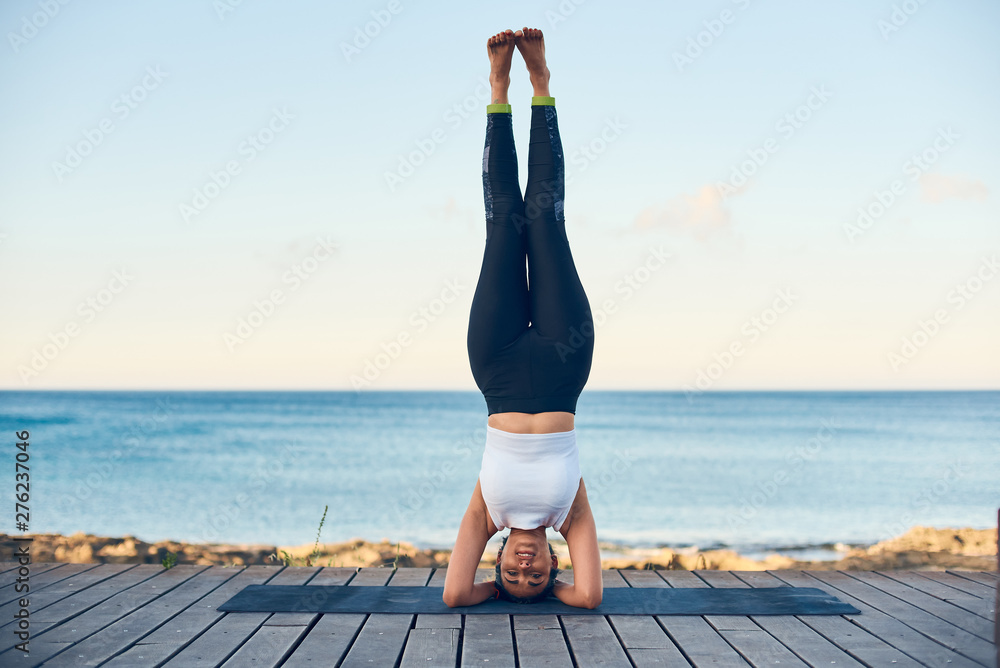 Beautiful fit woman practicing yoga against blue sea standing in headstand  pose. Stock Photo