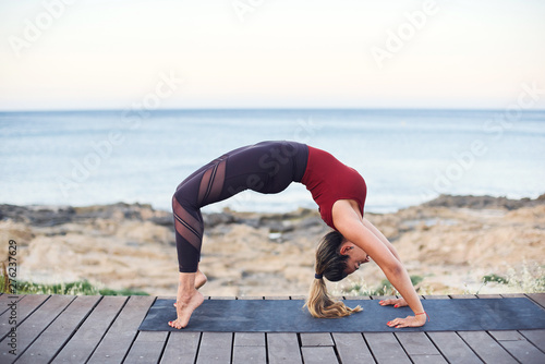 Young attractive woman practices yoga in wheel pose on the beach.