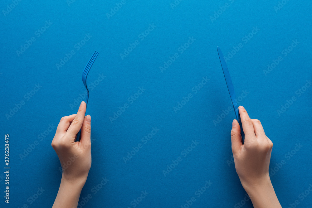 cropped view of woman holding blue plastic fork and knife on blue background - obrazy, fototapety, plakaty 