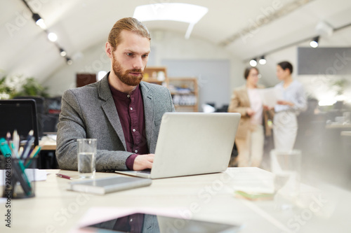 Portrait of handsome bearded businessman using laptop while working in open space office