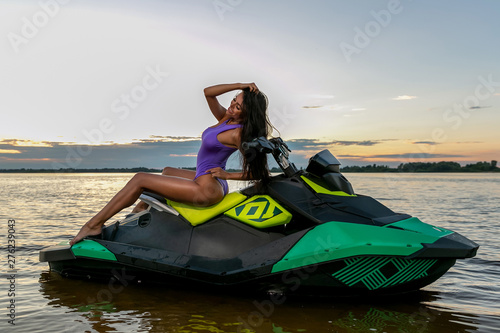Fotografie Young sexy girl in a blue swimsuit posing, sitting on a jet ski on the river in a sunshine