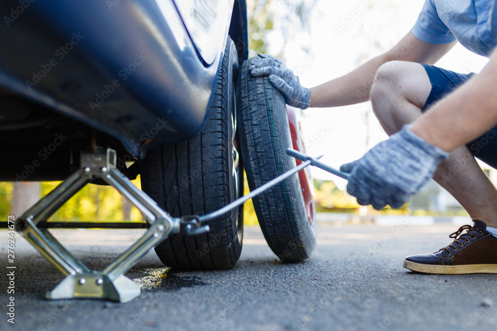 Obraz premium Mechanic in gloves lifts the car Jack on the side of the highway. Man is preparing to change the flat wheel lifting the car with a Jack on the side of the highway.