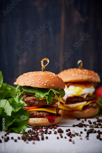 Two big classic american cheeseburger with two chops. Cheeseburgers on a linen tablecloth, decorated with different types of pepper and lettuce. Dark wooden background.