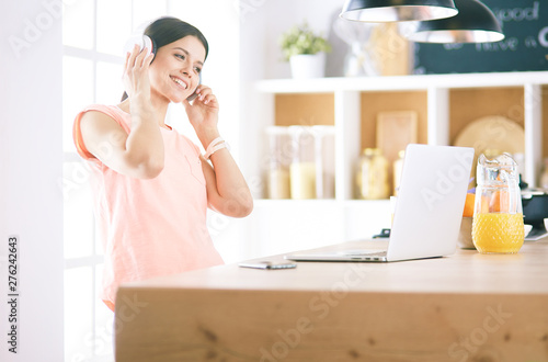 Portrait of a cheerful young woman listening to music with headphones and using laptop computer while standing at the kitchen