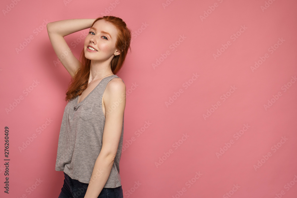 Redhead girl stands slightly sideways to the camera, touches the back of her head and smiles at the camera Redhead girl stands slightly sideways to the camera, touches the back of her head and smiles at the camera