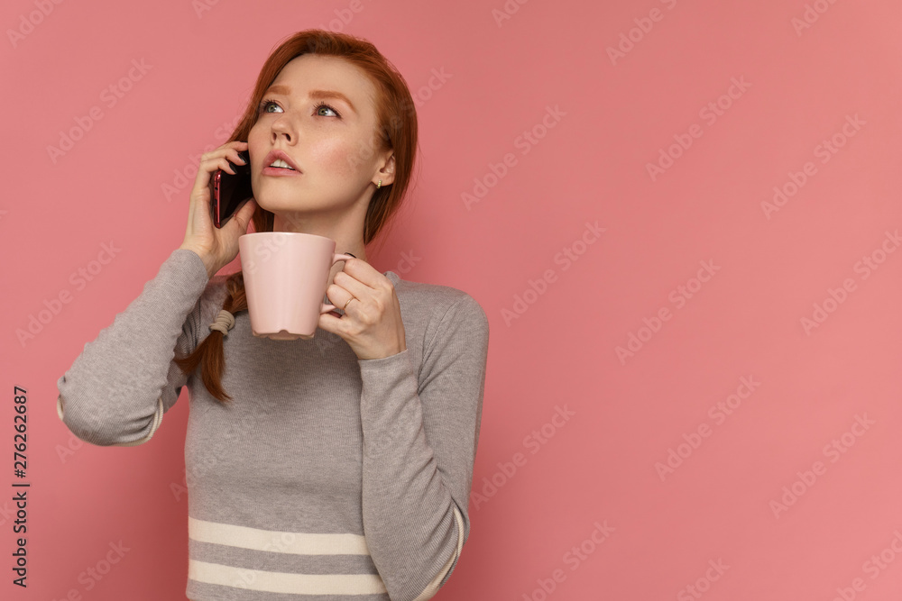 Redhead young woman talks on phone holding a cup in on one hand looking away Redhead young woman talks on phone holding a cup in on one hand looking away