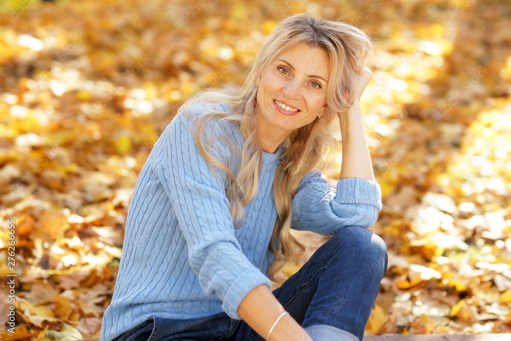 Happy beautiful blond woman sitting in park on sunny autumn day toned. Smyling woman in blue sweater outdoors on beautiful fall day Happy beautiful blond woman sitting in park on sunny autumn day toned. Smyling woman in blue sweater outdoors on beautiful fall day