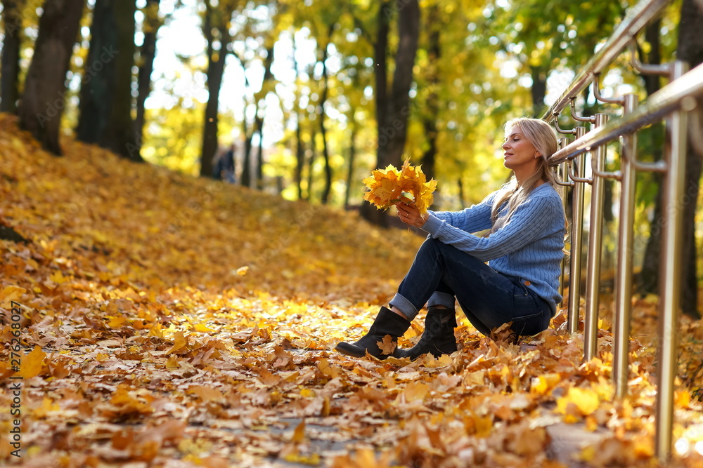 Happy beautiful blond woman sitting and holding colorful autumn leaves in park on sunny autumn day.