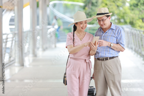 asian traveler couple husband and wife in pink and blue shirt with white nice hat and luggage walking in downtown with happy smiling face and using mobile for searching location during retirement age