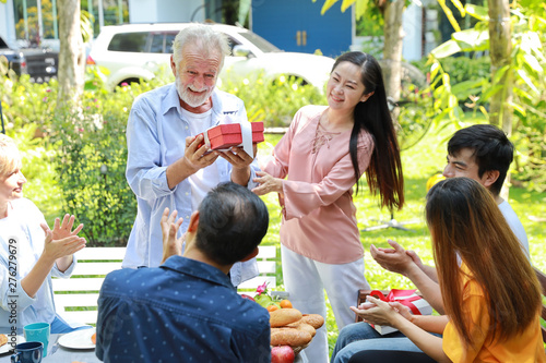 Happy multiethnic family giving surprise gift to caucasian grandfather on his happy birthday with happy smiling face in backyard outdoor on sunny day