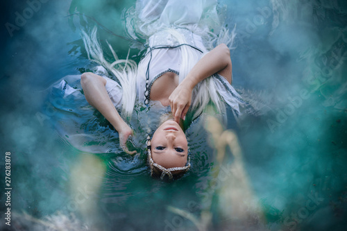 A girl elf in a white dress is floating in the water. Model in a medieval dress with pigtails floating in the lake