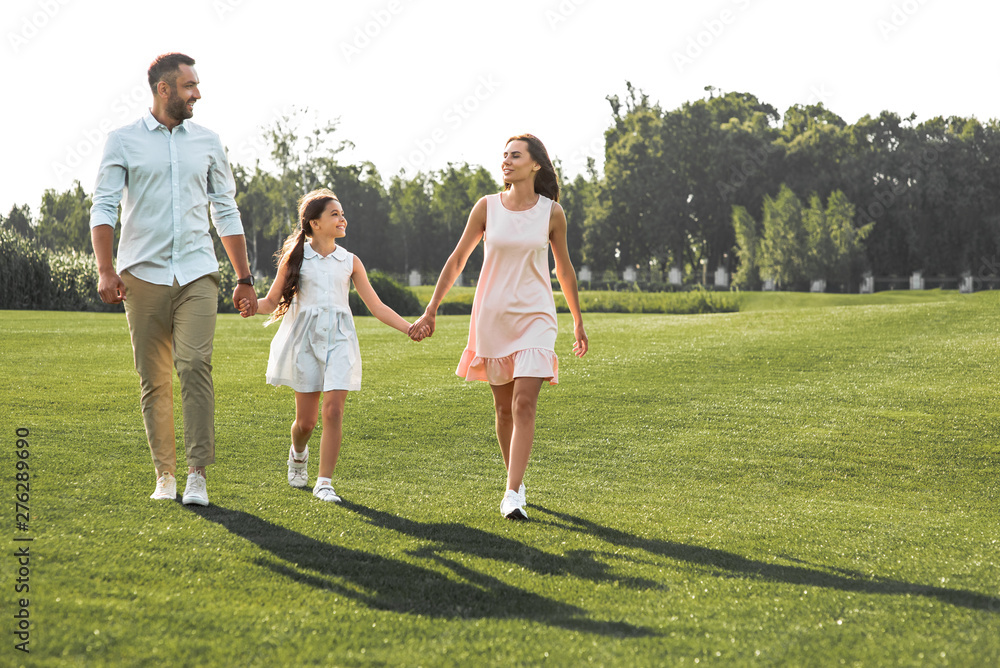 Enjoying time together. Full length of happy and young family of three holding hands, smiling and walking outdoors