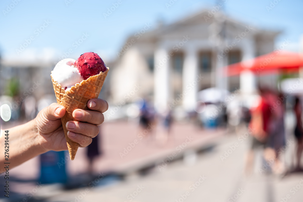 Woman's hand holding a waffle cone with 2 white and magenta ice cream balls on blurry old city background, sunny day light Woman's hand holding a waffle cone with 2 white and magenta ice cream balls on blurry old city background, sunny day light
