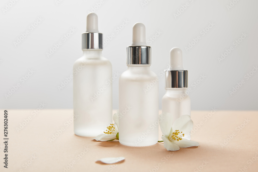 three cosmetic glass bottles with serum and few jasmine flowers with shadow on beige three cosmetic glass bottles with serum and few jasmine flowers with shadow on beige
