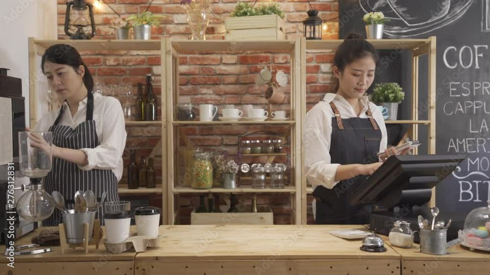 female shop assistant wearing apron using tablet after taking customer order in restaurant. two girls colleagues walking in counter working prepare coffee. small startup business owners partners. female shop assistant wearing apron using tablet after taking customer order in restaurant. two girls colleagues walking in counter working prepare coffee. small startup business owners partners.