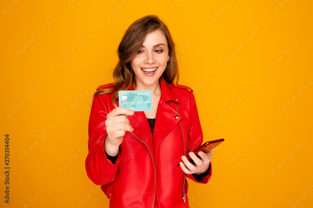Woman in the red jacket holding credit card and phone isolated over the yellow background Woman in the red jacket holding credit card and phone isolated over the yellow background