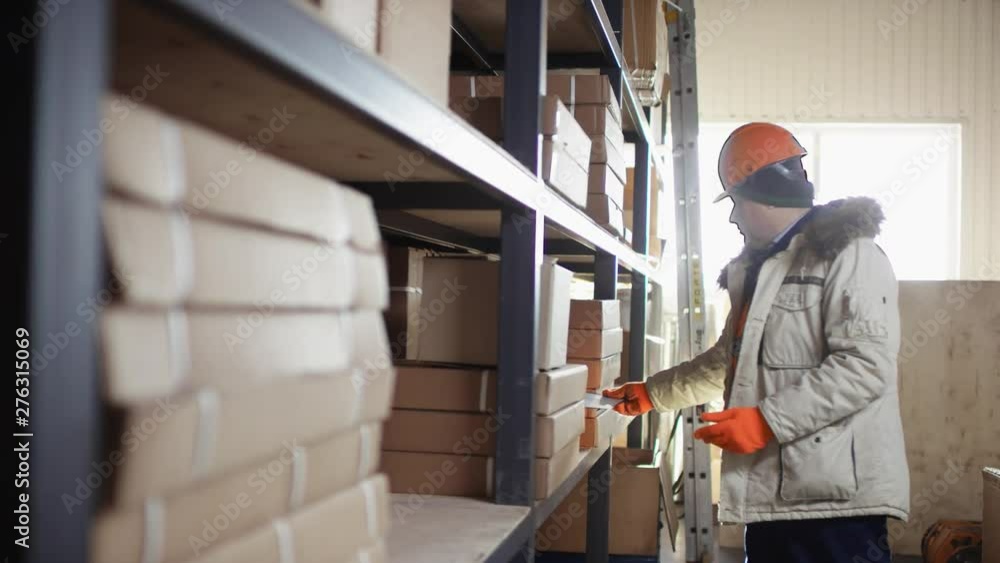 worker in a helmet and overalls stands next to the racks and remove box for right inventory with the tablet with flow camera worker in a helmet and overalls stands next to the racks and remove box for right inventory with the tablet with flow camera