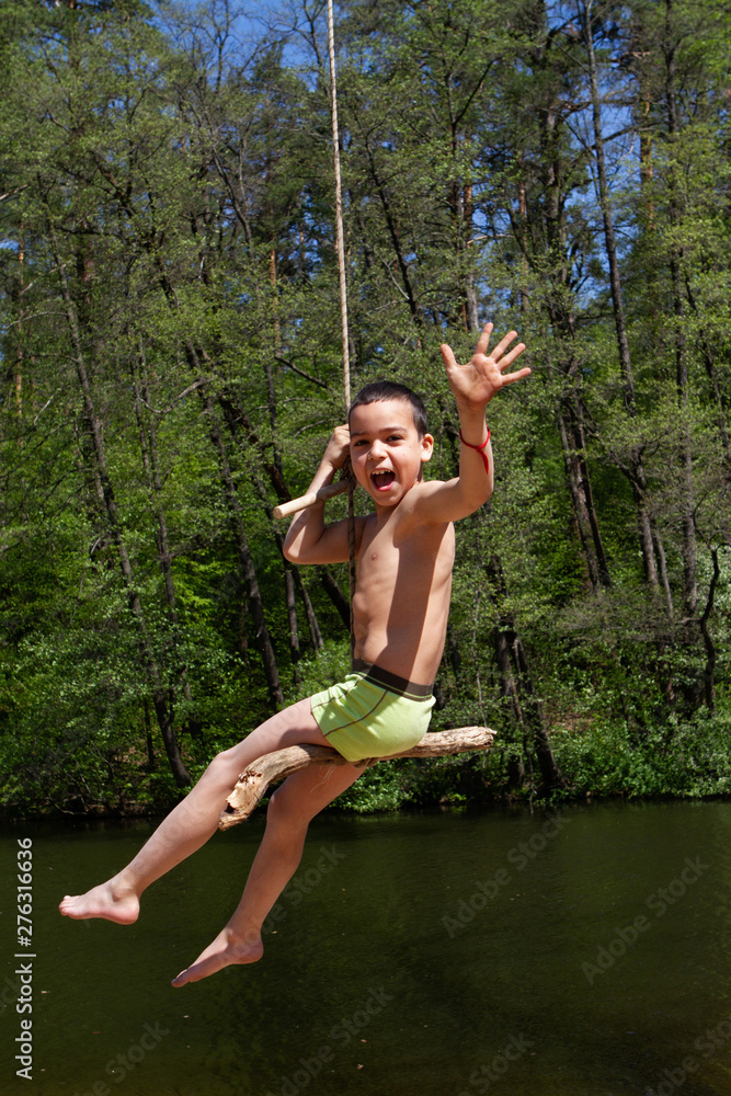 A little boy riding a rope swing in the forest above the surface of the lake.
