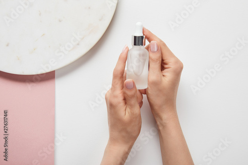 cropped view of cropped view of woman holding cosmetic glass bottle near plate on white pink surface
