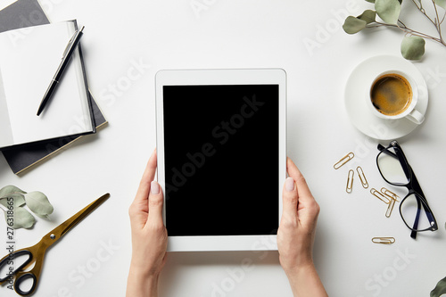 cropped view of woman holding digital tablet in hands near plants, stationery, scissors, coffee, glasses and notebooks with pen on white surface