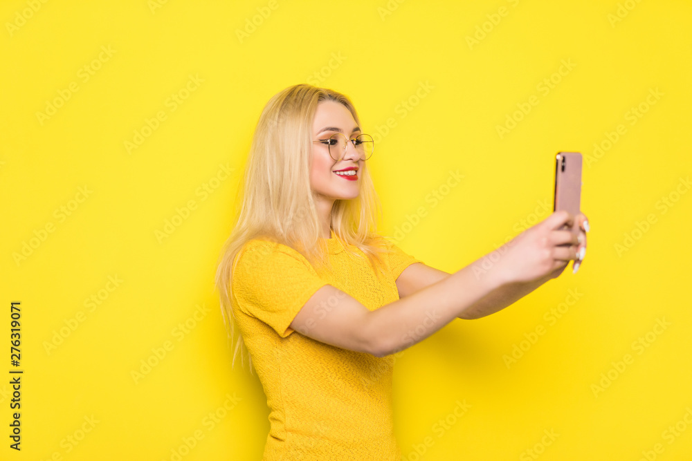 Portrait of cool cheerful girl having video-call with lover holding smart phone in hand shooting selfie on front camera isolated on yellow background Portrait of cool cheerful girl having video-call with lover holding smart phone in hand shooting selfie on front camera isolated on yellow background