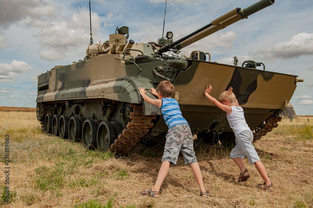 Two children in bright clothes are pushing away modern battle tank. Children play until tankists arrive. concept of contrasting innocent childhood and war