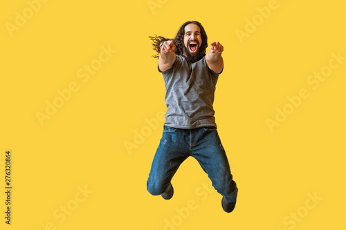 Portrait of surprised bearded young man with long curly hair in grey tshirt jumping, pointing and looking at camera with amazed happy funny face. indoor studio shot isolated on yellow background.