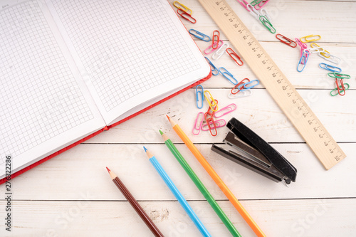 Various stationery placed on lumber table. From above colored pencils and open notebook arranged on wooden table near paper clips and stapler with ruler for studies