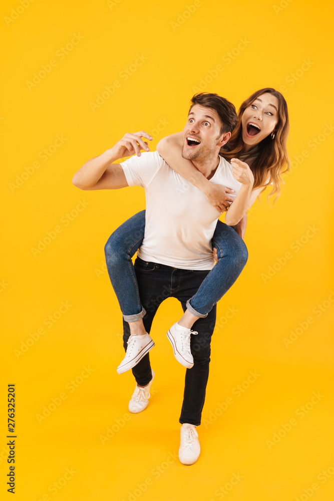 Full length portrait of pleased couple man and woman in basic t-shirts looking at copyspace together while doing piggyback ride isolated over yellow background Full length portrait of pleased couple man and woman in basic t-shirts looking at copyspace together while doing piggyback ride isolated over yellow background