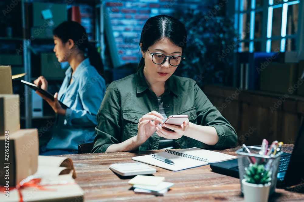 Fototapeta premium individual e commerce business at house studio. young hard working girl freelancer check orders and packing goods for delivery on mobile phone. employee colleague using tablet out time work night