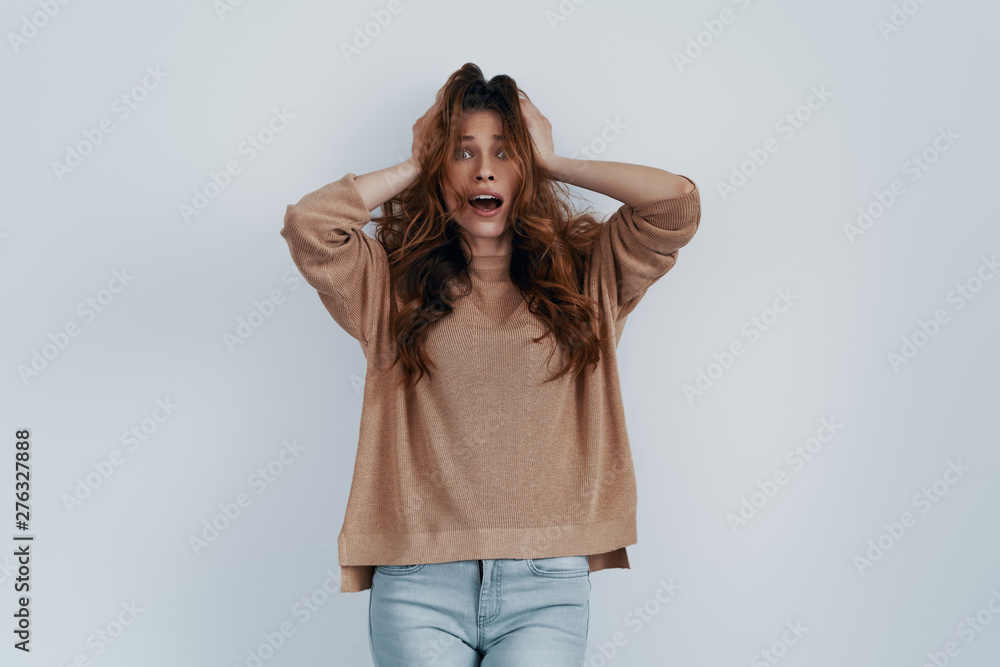 Feeling excited. Shocked young woman keeping hands in hair and smiling while standing against grey background Feeling excited. Shocked young woman keeping hands in hair and smiling while standing against grey background