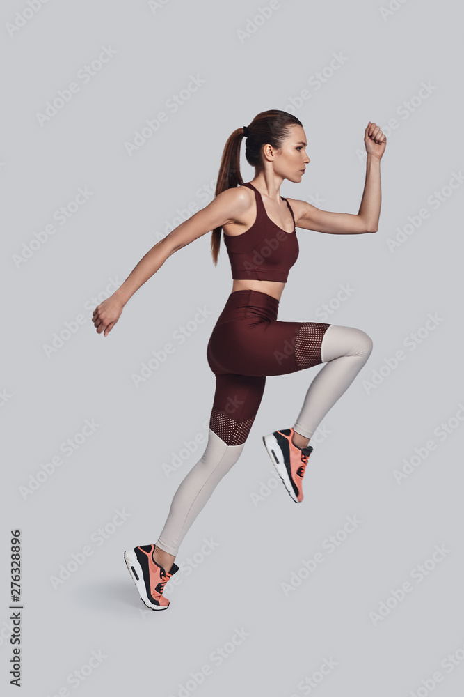 Always in good shape. Full length of attractive young woman in sports clothing jumping while exercising against grey background
