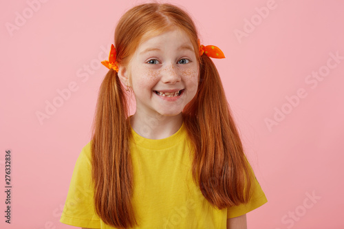 Close up of happy petite freckles red-haired girl with two tails, broadly smiling and looks cute, wears in yellow t-shirt, stands over pink background.